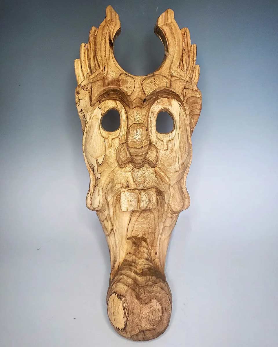 Wooden mask with abstract facial features, large eyes, prominent nose, open mouth, and horn-like structures on top against a light background.