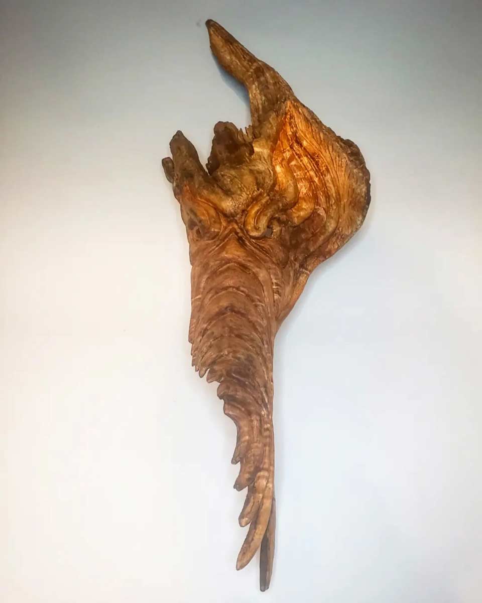 Intricately carved wooden sculpture resembling a mythical creature with a long, textured tail and horn-like protrusions on a light background.