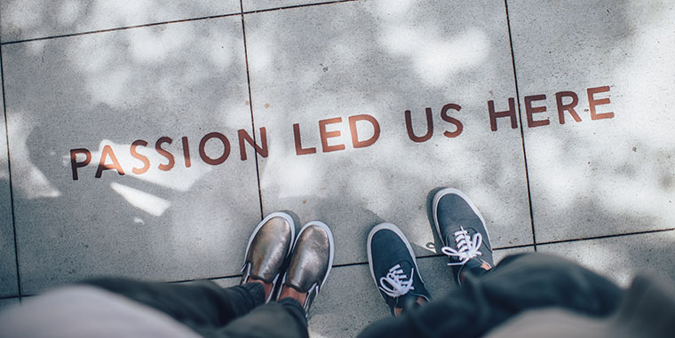 Two people only visible by their athletic sneakers standing on a concrete sideway which has a painted message that reads “Passion Led Us Here”
