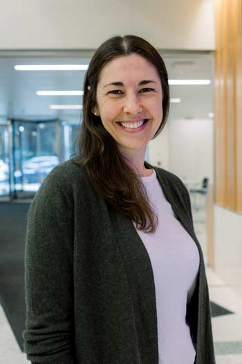 Person with long brown hair and green cardigan sweater smiling in hospital hallway