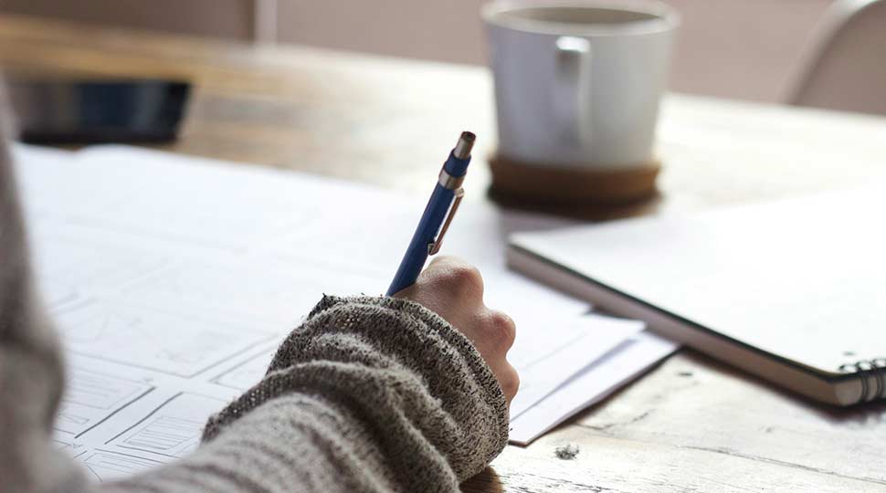 Person writing notes at table with pencil and mug in background.