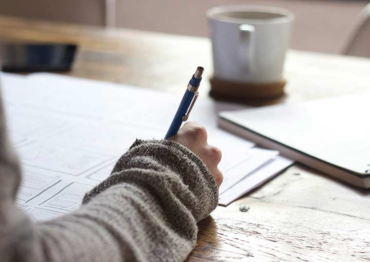 Person writing notes at table with pencil and mug in background.