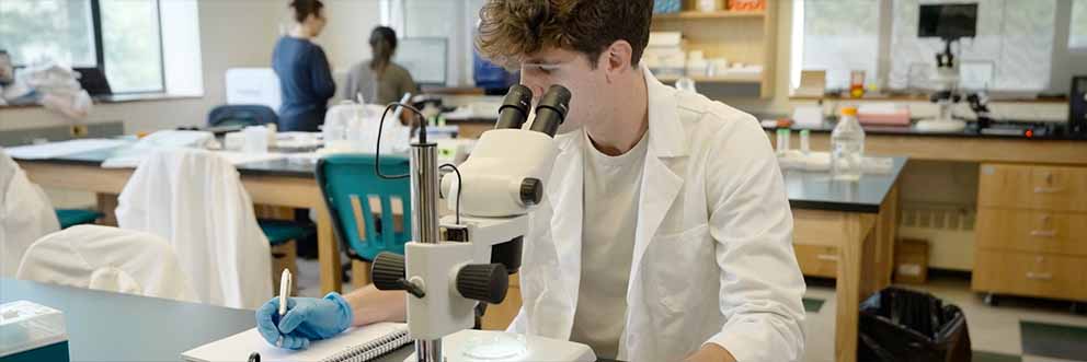 student in white lab coat seated at microscope