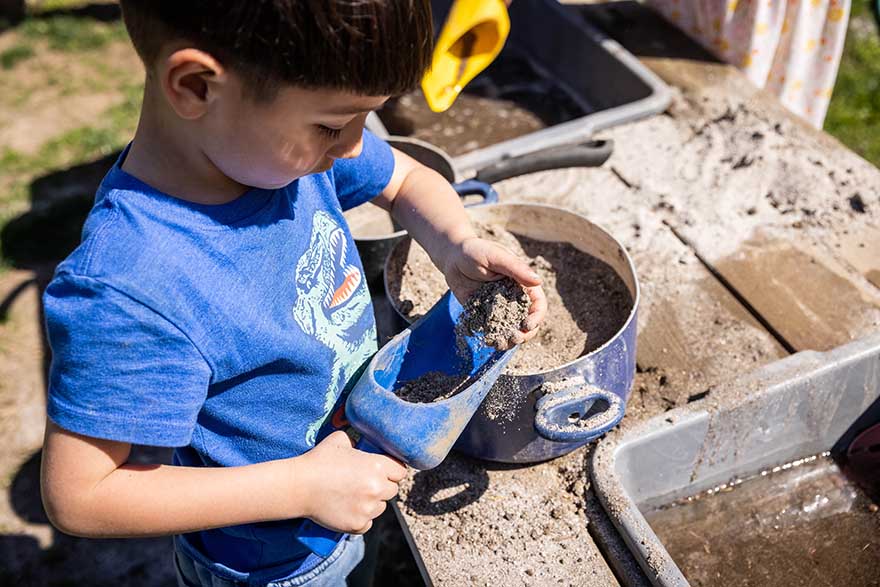 Young boy with dark hair uses toy shovel to scoop dirt into bucket  