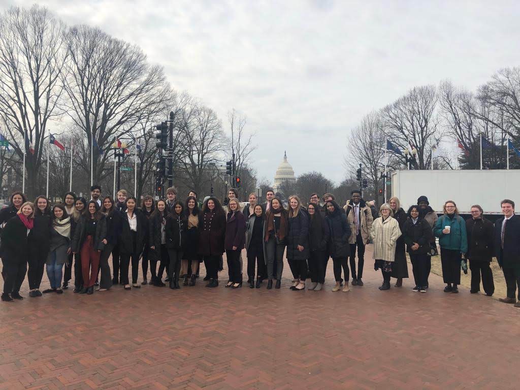 A large group of students stand, wearing coats in the cold weather, with the round top of the Capitol building visible between bare tree branches behind them.