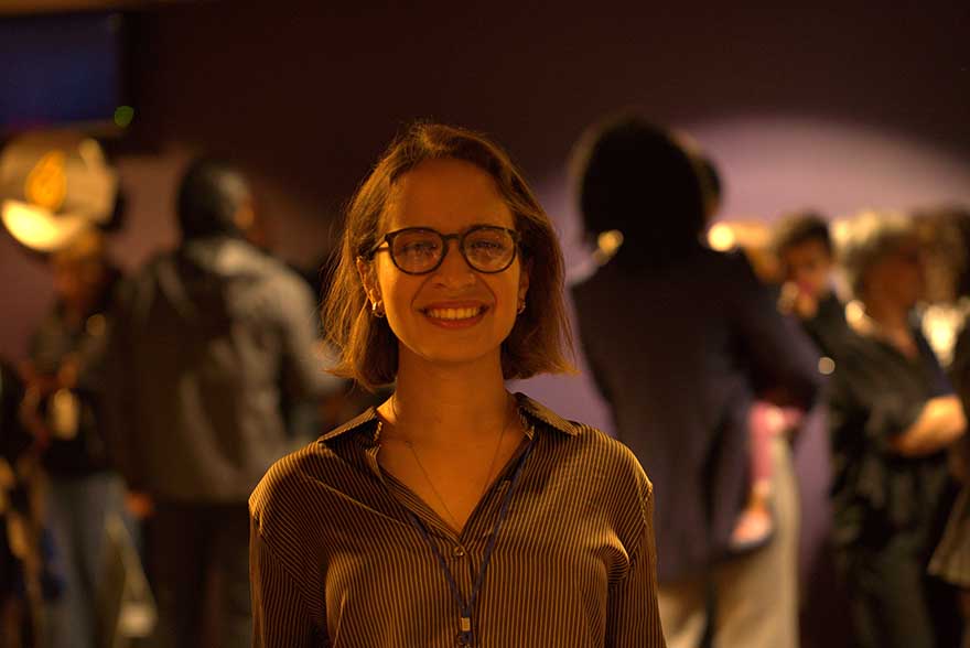 Young woman with glasses looking at camera with out of focus people behind her.