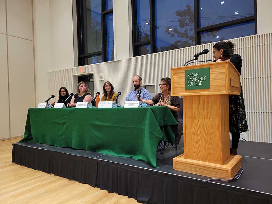 4 women and 1 man sit behind table on a stage. 1 woman stands at a lectern on stage.
