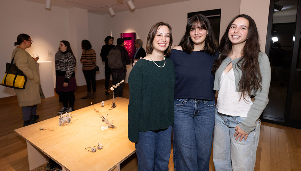 Three students pose in front of artwork and smile at the camera