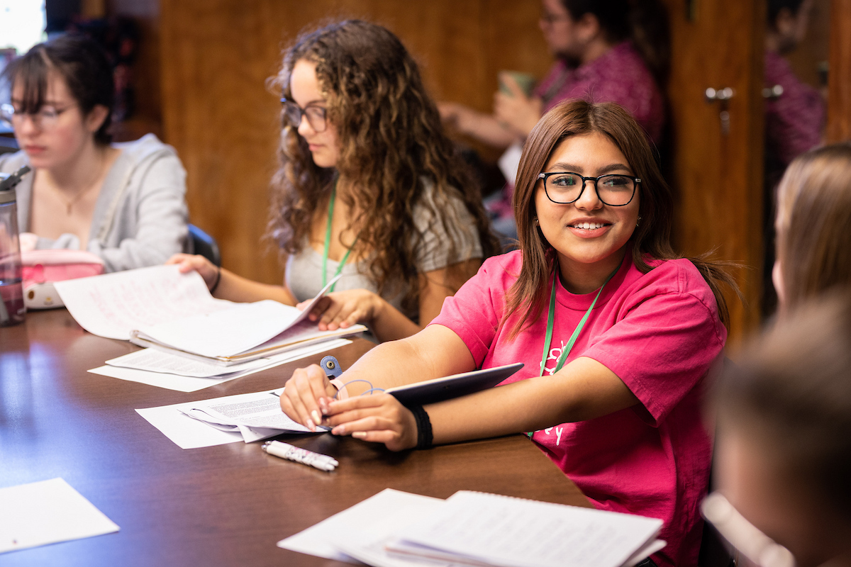 Student smiling at someone as they sit around a classroom table surrounded by others
