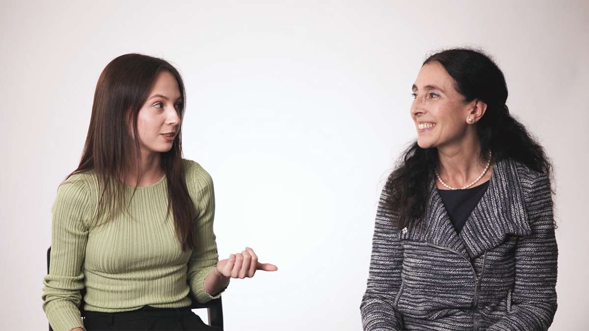 Two women seated engaged in conversation against a plain studio background.