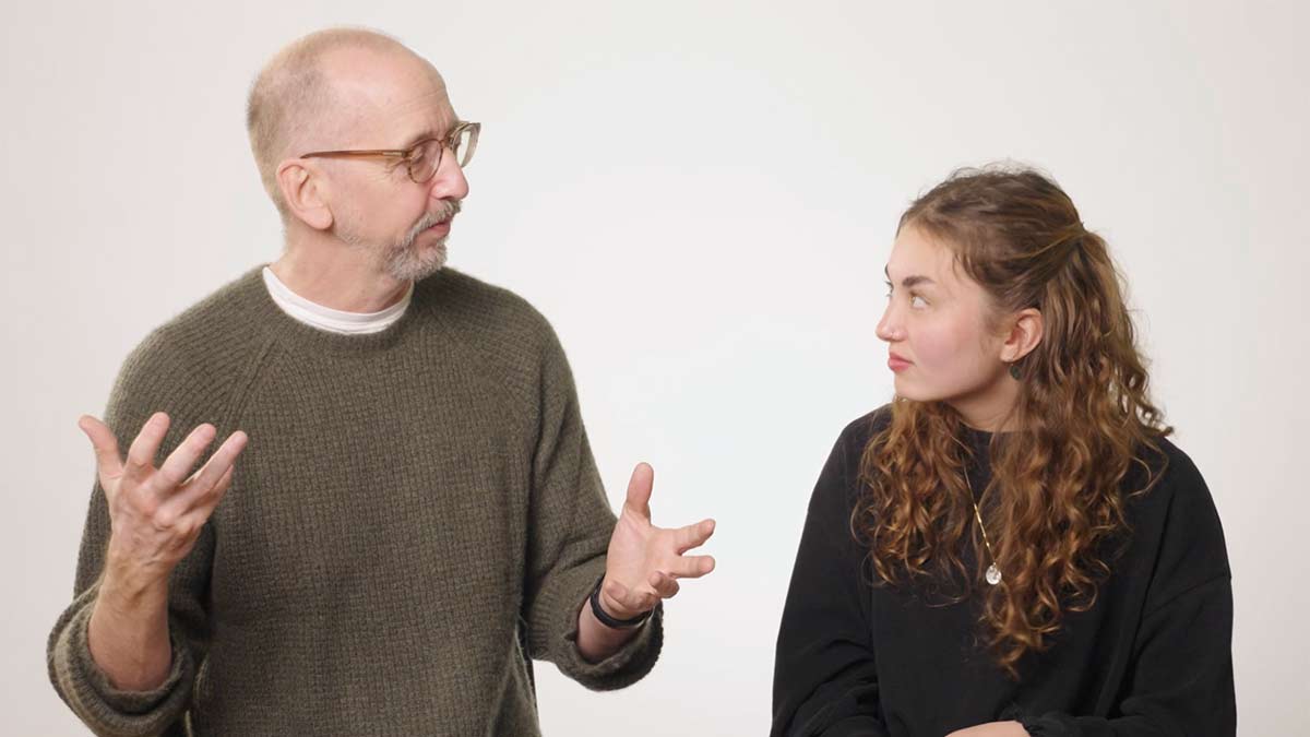 A man gestures while speaking to a woman seated beside him, both engaged in conversation against a plain studio background.