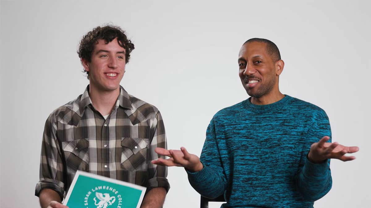 Two men sit side by side against a plain light background, smiling at the camera.