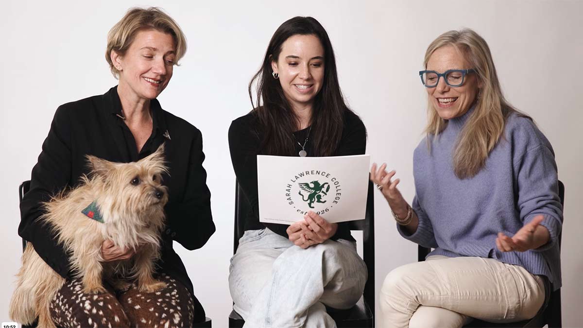 Three women sitting and smiling in conversation against a plain studio background. One woman holding a dog.