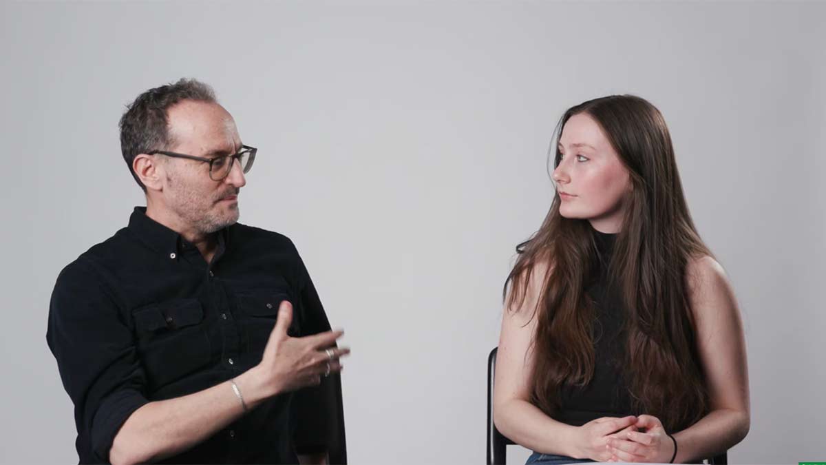 A man gestures while speaking to a woman seated beside him, both engaged in conversation against a plain studio background.