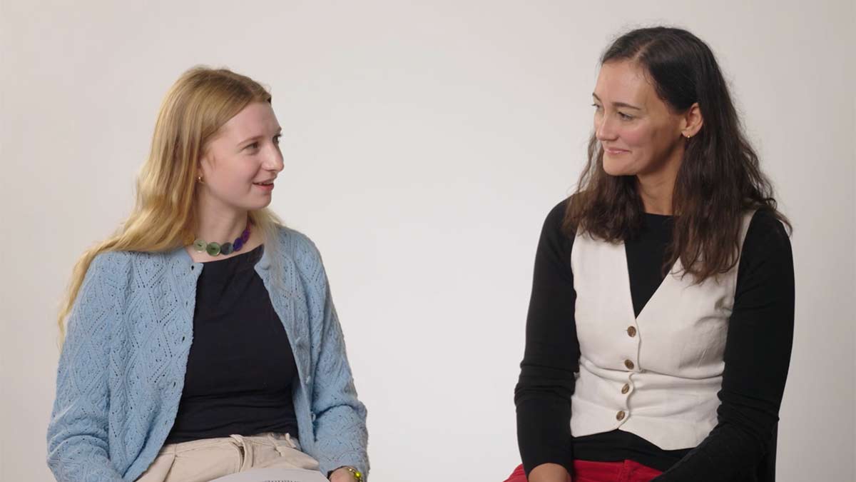 Two women seated engaged in conversation against a plain studio background.