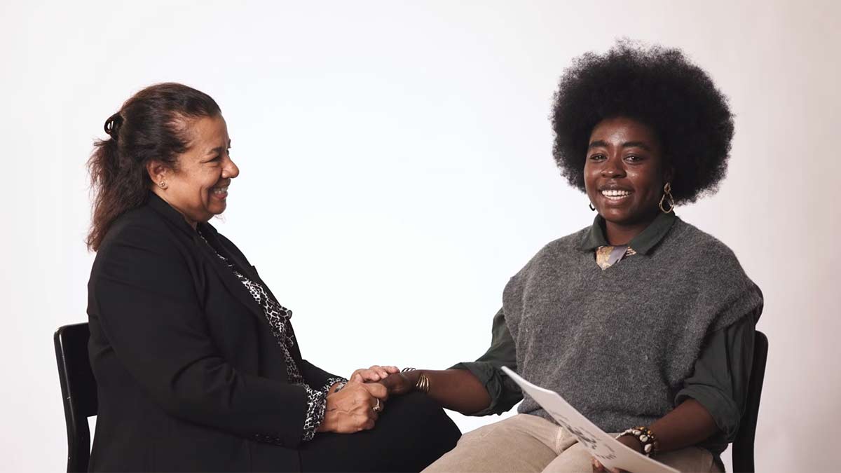 Two women seated and smiling at each other while holding hands, suggesting a warm, supportive conversation in a studio setting.
