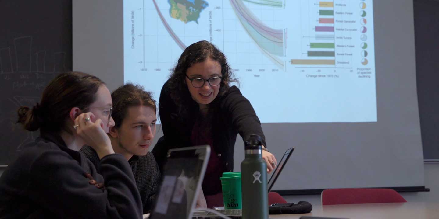 Projection screen showing charts and data in the background. Teacher with two students discussing a project.
