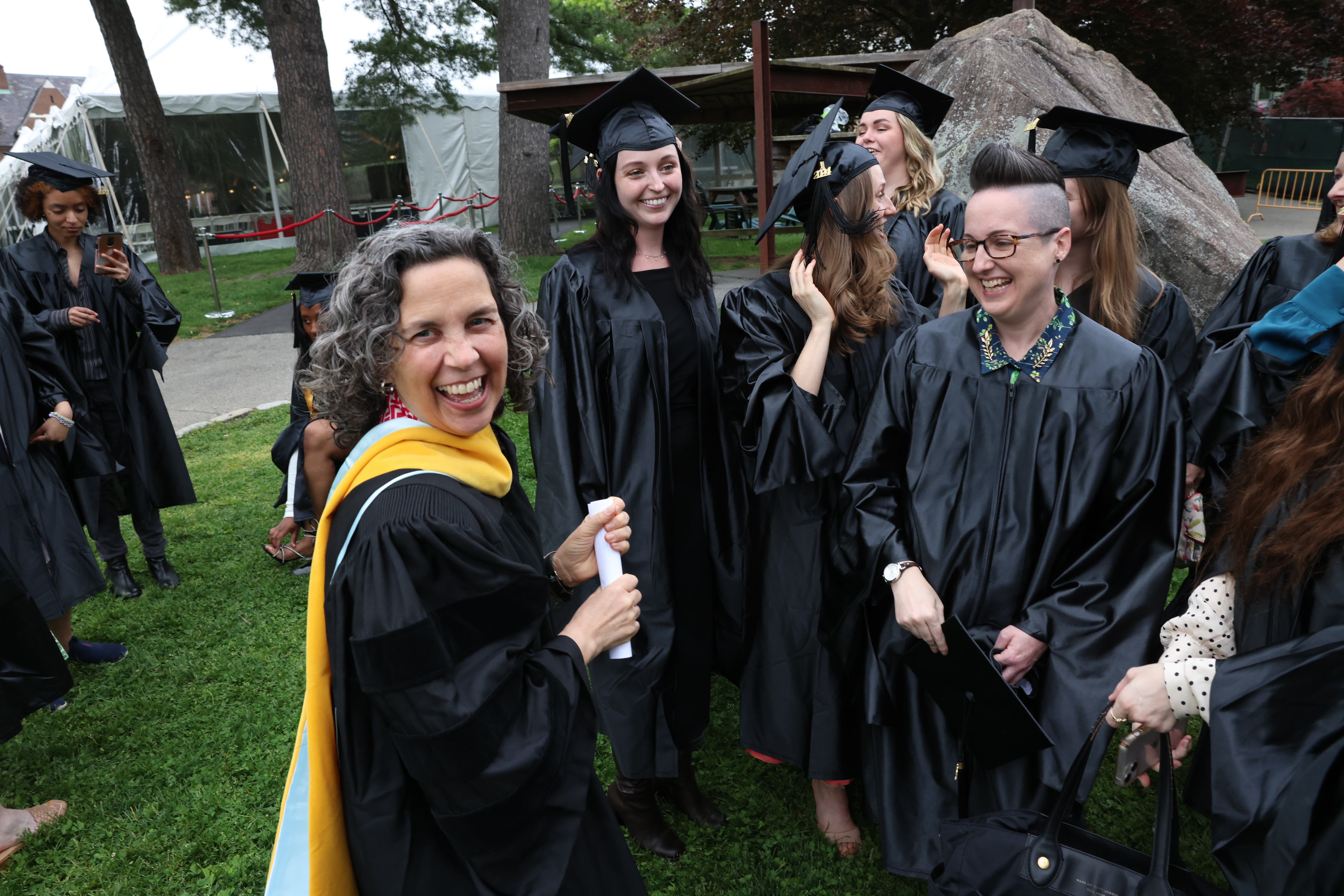People in graduation gowns and caps standing on a lawn, smiling and chatting before a ceremony. Trees and a large rock are in the background.